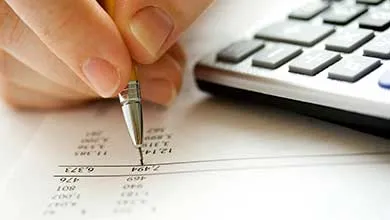 Hand holding mechanical pencil over spreadsheet; calculator in background.